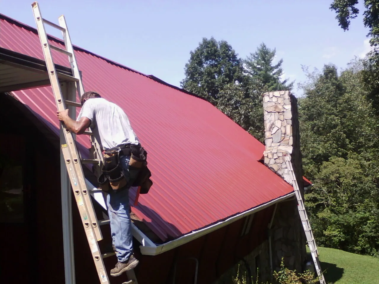 Roofer on a ladder beside a red metal roof and stone chimney — Soffit & Fascia Repair by Roofing Brothers in Tangent